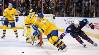 Erik Brännström i gul svensk tröja driver pucken och jagas av en amerikansk spelare. i bakgrunden syns den svenska målvakten och en annan svensk back.